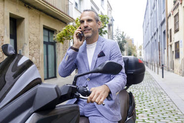 Mature businessman talking over mobile phone sitting on motorcycle - RFTF00361