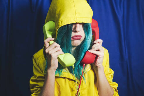 Introvert woman in hooded shirt talking on green and red telephones in front of blue backdrop - SVCF00277