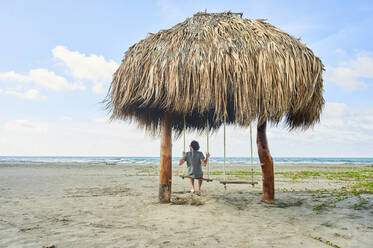 Mann sitzt auf einer Schaukel unter einer Hütte mit Strohdach am Strand, Cartagena de Indias, Kolumbien - KIJF04510