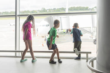 Schwester mit Brüdern, die vom Fenster des Flughafenterminals aus die Vorbereitung des Flugzeugs zum Einsteigen beobachten - OSF01305