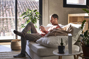 Man using mobile phone sitting on sofa at home - VEGF06140