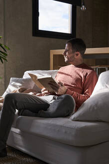 Nachdenklicher Mann sitzt mit einem Buch auf dem Sofa zu Hause - VEGF06149