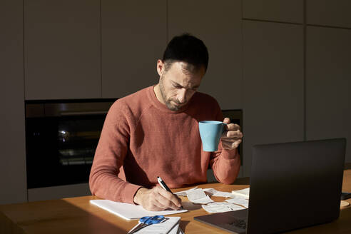 Man with coffee cup writing on paper at desk - VEGF06176