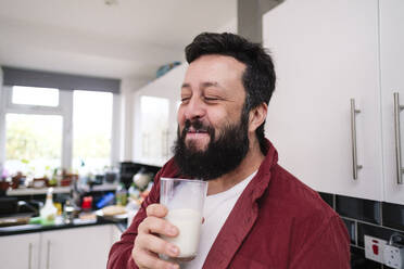 Lächelnder Mann genießt ein Glas Milch in der Küche zu Hause - ASGF03232