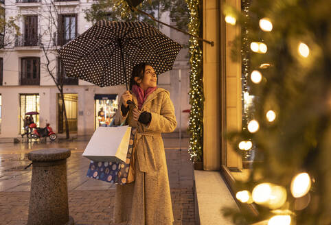 Glückliche junge Frau mit Regenschirm beim Schaufensterbummel auf dem Fußweg - JCCMF08728