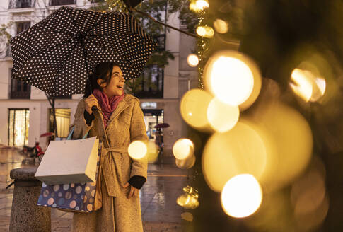 Glückliche Frau mit Regenschirm beim Schaufensterbummel auf dem Fußweg - JCCMF08729