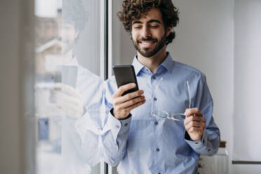 Lächelnder Geschäftsmann, der ein Smartphone benutzt und eine Brille im Büro hält - EBBF07551