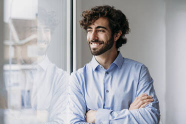 Happy contemplative businessman looking through window in office - EBBF07554