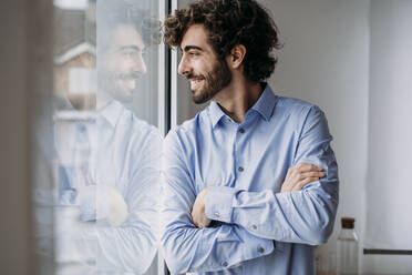 Happy businessman standing with arms crossed looking through window - EBBF07555