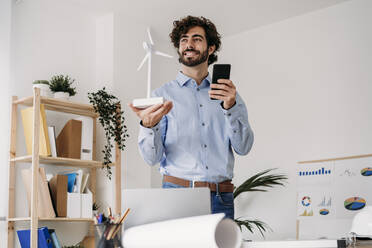 Nachdenklicher Ingenieur mit Windrad und Mobiltelefon im Büro - EBBF07575