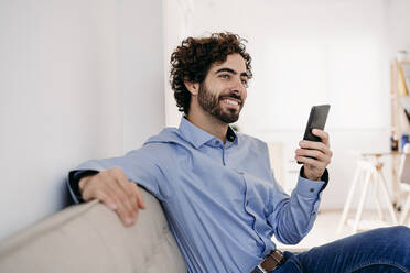 Smiling businessman sitting with smart phone in office - EBBF07583