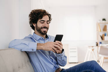 Happy young businessman using smart phone sitting on sofa in office - EBBF07585