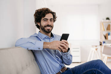 Happy young businessman sitting with smart phone on sofa at office - EBBF07586