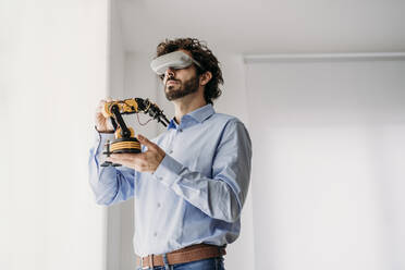 Geschäftsmann mit VR-Brille hält Roboterarm im Büro - EBBF07591