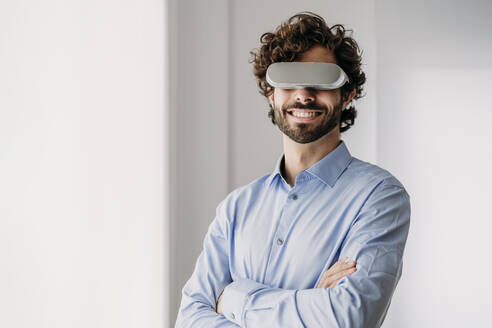 Lächelnder Ingenieur mit Virtual-Reality-Brille, der mit verschränkten Armen im Büro steht - EBBF07594