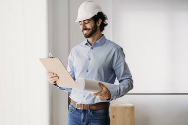 Smiling architect holding blueprints by window in office - EBBF07601