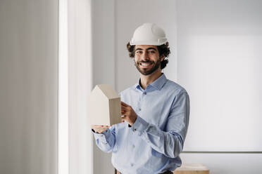 Glücklicher Architekt hält Hausmodell in der Nähe des Fensters im Büro - EBBF07607