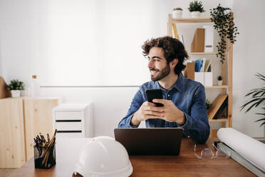 Glücklich lächelnder Ingenieur, der mit Smartphone und Laptop auf dem Schreibtisch im Büro sitzt - EBBF07620