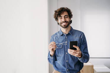 Happy young entrepreneur with smart phone standing by window in office - EBBF07631