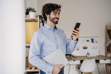 Glücklicher junger Ingenieur, der ein Mobiltelefon benutzt und ein Smartphone im Büro hält - EBBF07633