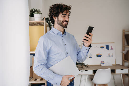 Happy young engineer using mobile phone and holding smart phone at office - EBBF07633