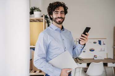 Lächelnder junger Ingenieur mit Smartphone und Laptop im Büro - EBBF07634