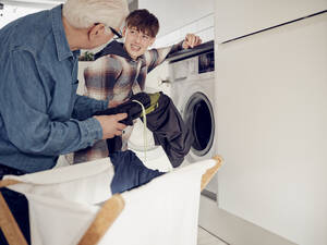 Grandson and grandfather putting laundry into washing machine at home - PWF00408
