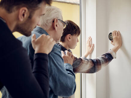 Sohn, Vater und Großvater betrachten den intelligenten Thermostat an der Wand in der Küche - PWF00411