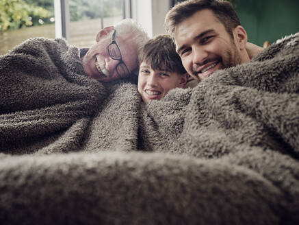 Glücklicher Großvater, Vater und Sohn kuscheln unter einer Decke auf der Couch im Wohnzimmer - PWF00430