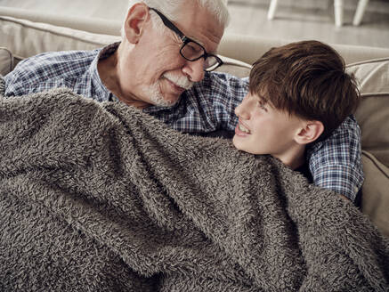 Grandfather and son snuggling under a blanket on couch in living room - PWF00432