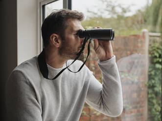 Man looking out of the window at home with binoculars - PWF00438