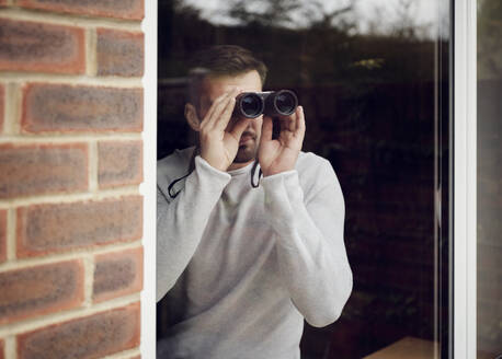 Mann schaut zu Hause mit Fernglas aus dem Fenster - PWF00439
