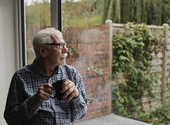 Nachdenklicher älterer Mann mit einer Tasse Kaffee, der zu Hause aus dem Fenster schaut - PWF00443