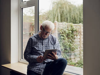 Älterer Mann am Fenster zu Hause mit digitalem Tablet - PWF00444