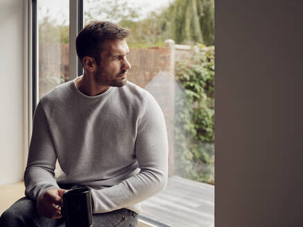 Pensive man with cup of coffee looking out of the window at home - PWF00445