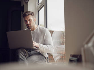 Mann am Fenster zu Hause mit Laptop - PWF00447