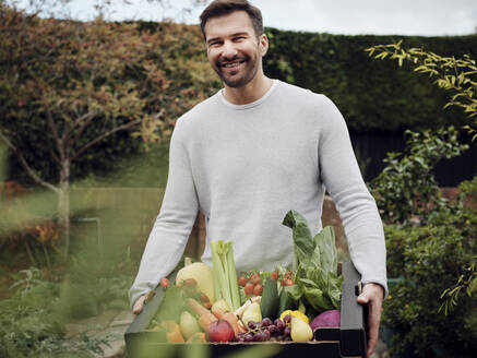 Glücklicher Mann trägt eine Kiste mit frischem Gemüse im Garten - PWF00452