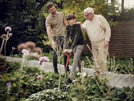 Father and grandfather watching teenage boy digging in garden - PWF00472