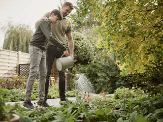 Father and son standing in garden watering plants - PWF00477