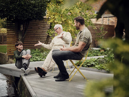 Vater, Sohn und Enkel sitzen am Gartenteich, unterhalten sich und entspannen sich - PWF00480