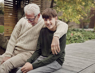 Grandson and grandfather sitting in garden talking together - PWF00486