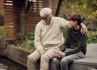 Grandson and grandfather sitting in garden talking together - PWF00488