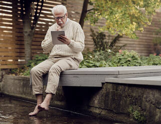 Senior man sitting by garden pond using digital tablet - PWF00492