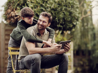 Sohn umarmt Vater im Garten sitzend mit digitalem Tablet - PWF00494