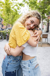 Lächelnde junge Frau mit blondem Haar, die eine Freundin auf dem Fußweg umarmt - WPEF06865