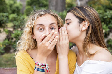 Woman whispering into shocked friend's ear in park - WPEF06887
