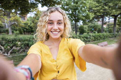 Glückliche junge Frau mit blondem Haar macht Selfie im Park - WPEF06889