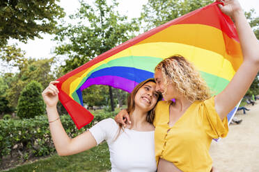 Happy lesbian couple holding rainbow flag embracing each other in park - WPEF06903