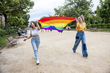 Lächelndes lesbisches junges Paar mit Regenbogenfahne im Park - WPEF06904