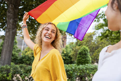 Happy woman holding rainbow flag looking at girlfriend in park - WPEF06905